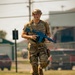 A military or law enforcement personnel in full tactical gear is running across a grassy field while carrying a bright blue training weapon. The individual is wearing OCP (Operational Camouflage Pattern) uniform, a tan tactical helmet, and a plate carrier vest. The blue-colored firearm indicates this is a training exercise using a simulated or inert weapon replica.