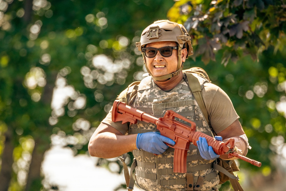 A person wearing military-style tactical gear, including a camouflage vest, tan helmet, and sunglasses, is holding a red/orange training rifle replica. The individual is smiling and appears to be in an outdoor setting with green foliage in the background. The red-colored firearm is a standard indicator of a training or inert dummy weapon used in military or law enforcement exercises.