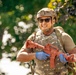 A person wearing military-style tactical gear, including a camouflage vest, tan helmet, and sunglasses, is holding a red/orange training rifle replica. The individual is smiling and appears to be in an outdoor setting with green foliage in the background. The red-colored firearm is a standard indicator of a training or inert dummy weapon used in military or law enforcement exercises.