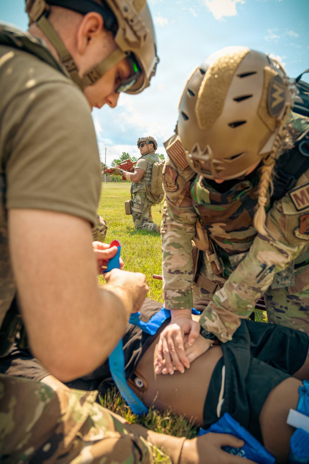 Military personnel in full combat gear and helmets are performing tactical combat casualty care (TCCC) on a simulated casualty lying on the ground. Two operators are actively treating the patient while a third stands in the background appearing to observe or coordinate. The scene appears to be a training exercise conducted outdoors on a grassy field.