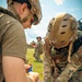 Military personnel in full combat gear and helmets are performing tactical combat casualty care (TCCC) on a simulated casualty lying on the ground. Two operators are actively treating the patient while a third stands in the background appearing to observe or coordinate. The scene appears to be a training exercise conducted outdoors on a grassy field.