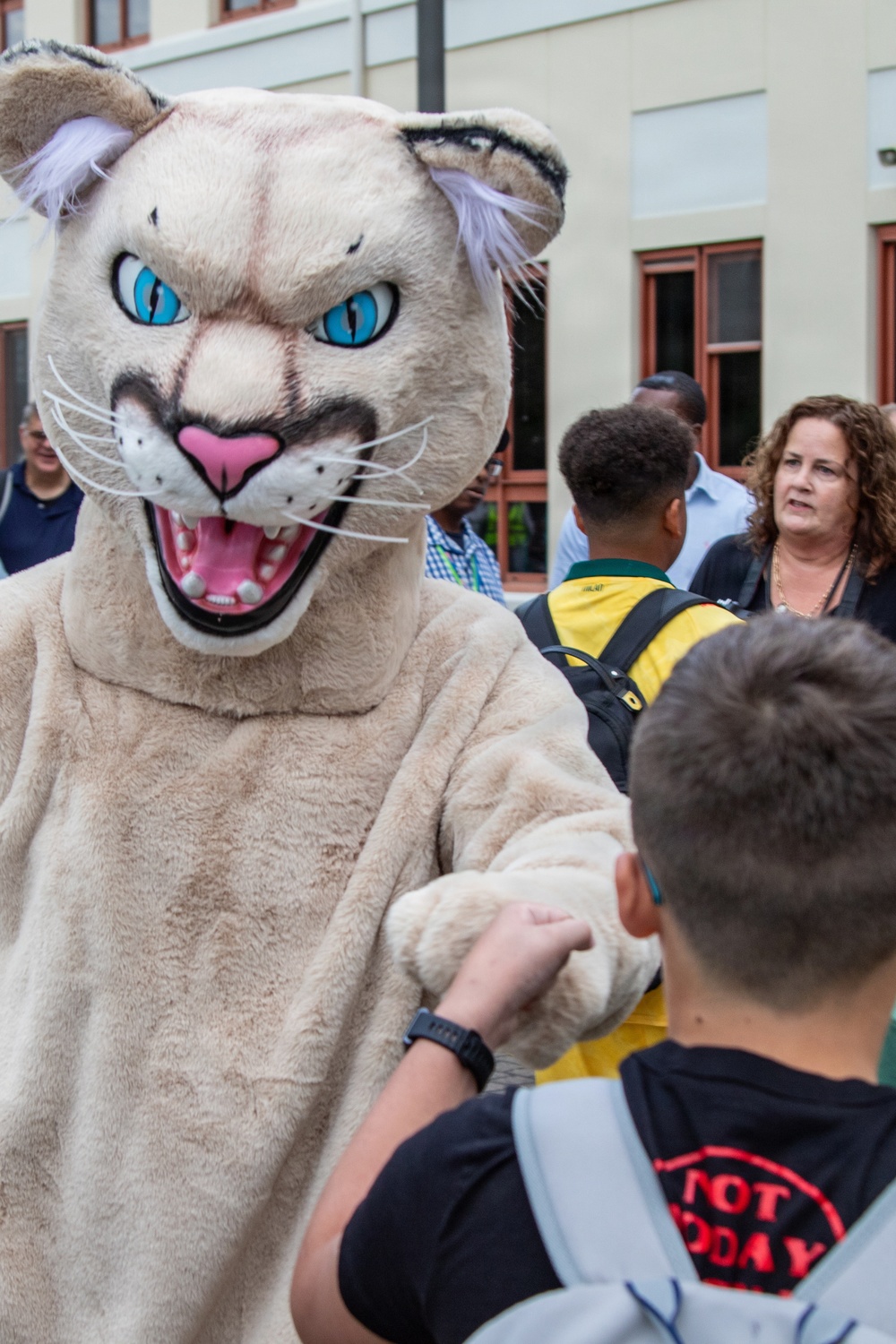 A person wearing a large wildcat or cougar mascot costume with blue eyes and an open mouth is interacting with a young boy who is shaking its hand/paw. The scene appears to take place outdoors near a building, likely at a school or community event. Several adults and another youth are visible in the background.