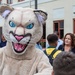 A person wearing a large wildcat or cougar mascot costume with blue eyes and an open mouth is interacting with a young boy who is shaking its hand/paw. The scene appears to take place outdoors near a building, likely at a school or community event. Several adults and another youth are visible in the background.