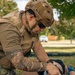 A military or tactical personnel wearing full combat gear — including a tan helmet, plate carrier, and camouflage pants — is applying a tourniquet to a casualty's leg on a grassy outdoor area. The casualty's lower leg displays simulated traumatic wound makeup consistent with a training exercise scenario. Medical supplies including bandages and blue gloves are visible on the ground nearby.