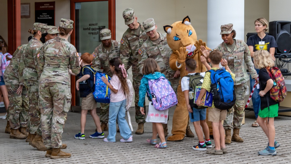 A group of U.S. Army soldiers in camouflage uniforms are greeting young children with backpacks outside what appears to be Vicenza Elementary School. A school mascot costume resembling a cougar or wildcat is present and high-fiving students. The scene appears to be a back-to-school welcome event at a military base school.