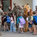 A group of U.S. Army soldiers in camouflage uniforms are greeting young children with backpacks outside what appears to be Vicenza Elementary School. A school mascot costume resembling a cougar or wildcat is present and high-fiving students. The scene appears to be a back-to-school welcome event at a military base school.