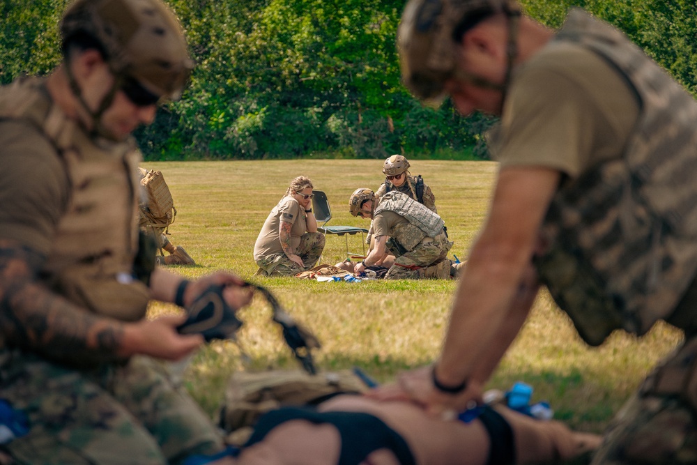 Military personnel are conducting tactical combat casualty care (TCCC) or field medical training exercises on an open grass field. Multiple teams are simultaneously practicing trauma treatment on simulated casualties lying on the ground. The setting appears to be a daytime outdoor training exercise with personnel in various military uniforms and gear.