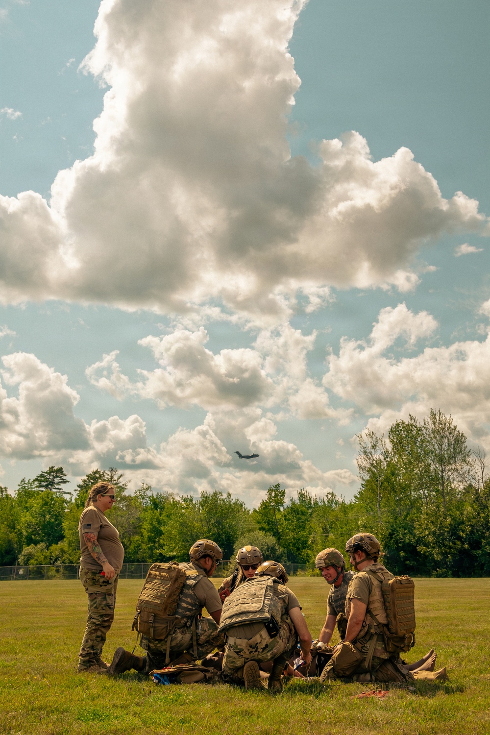 A group of military personnel in camouflage uniforms and tactical gear are conducting what appears to be a combat casualty care or tactical combat medicine training exercise on an open grass field. Several soldiers are kneeling around a simulated casualty on the ground while one individual stands observing, likely acting as an instructor or evaluator. A military transport aircraft is visible in the background sky, and the setting appears to be a training installation surrounded by trees.