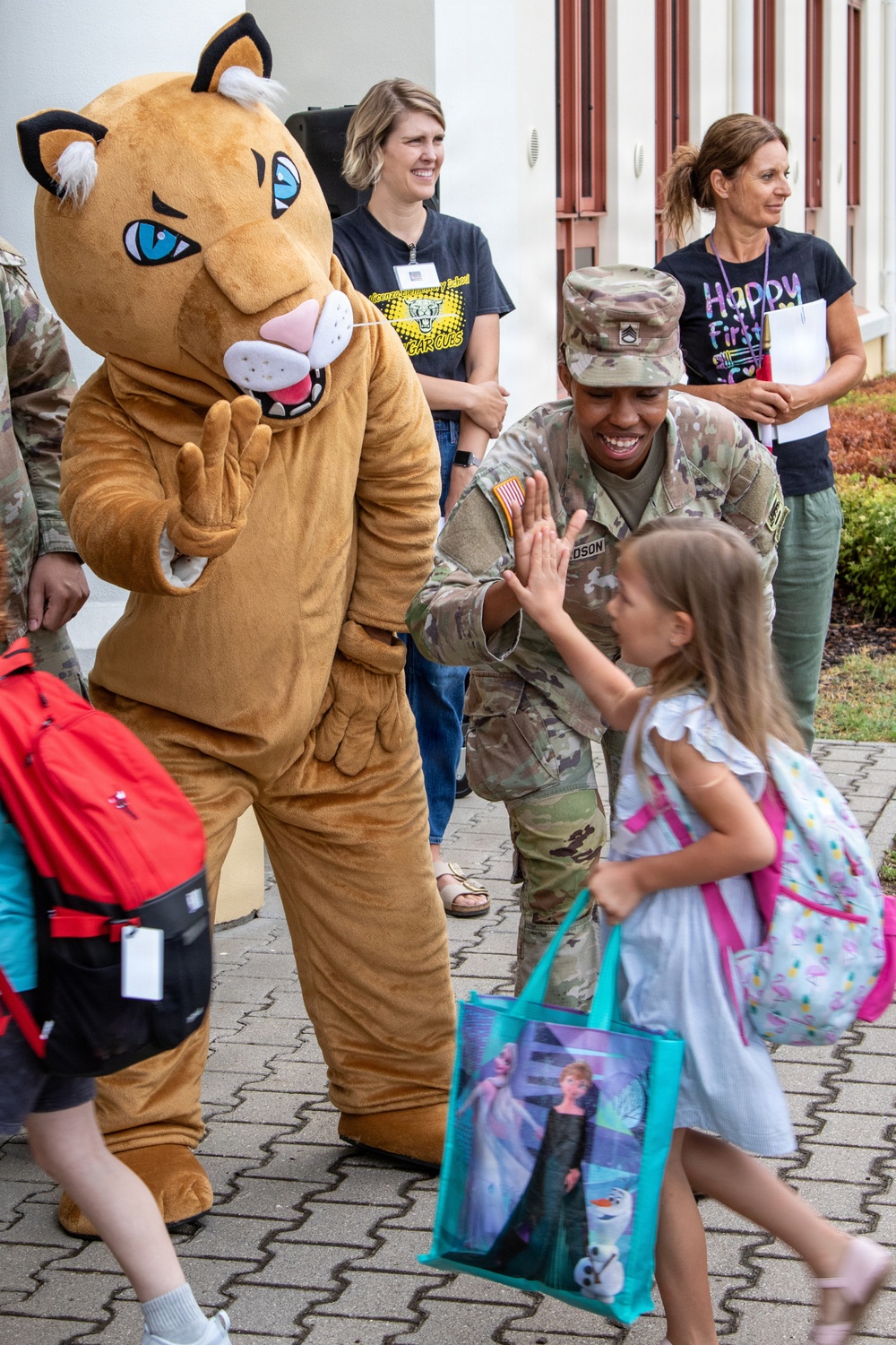 A U.S. Army soldier in Army Combat Uniform (ACU/OCP pattern) gives a high-five to a young girl carrying a Frozen-themed tote bag and a floral backpack. A large cougar/puma mascot costume stands nearby, suggesting this is a school welcome event. Two adult women in the background, one wearing a 'Happy First Day' shirt, observe the interaction, indicating this is likely a first day of school celebration at a military installation school.