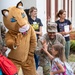 A U.S. Army soldier in Army Combat Uniform (ACU/OCP pattern) gives a high-five to a young girl carrying a Frozen-themed tote bag and a floral backpack. A large cougar/puma mascot costume stands nearby, suggesting this is a school welcome event. Two adult women in the background, one wearing a 'Happy First Day' shirt, observe the interaction, indicating this is likely a first day of school celebration at a military installation school.