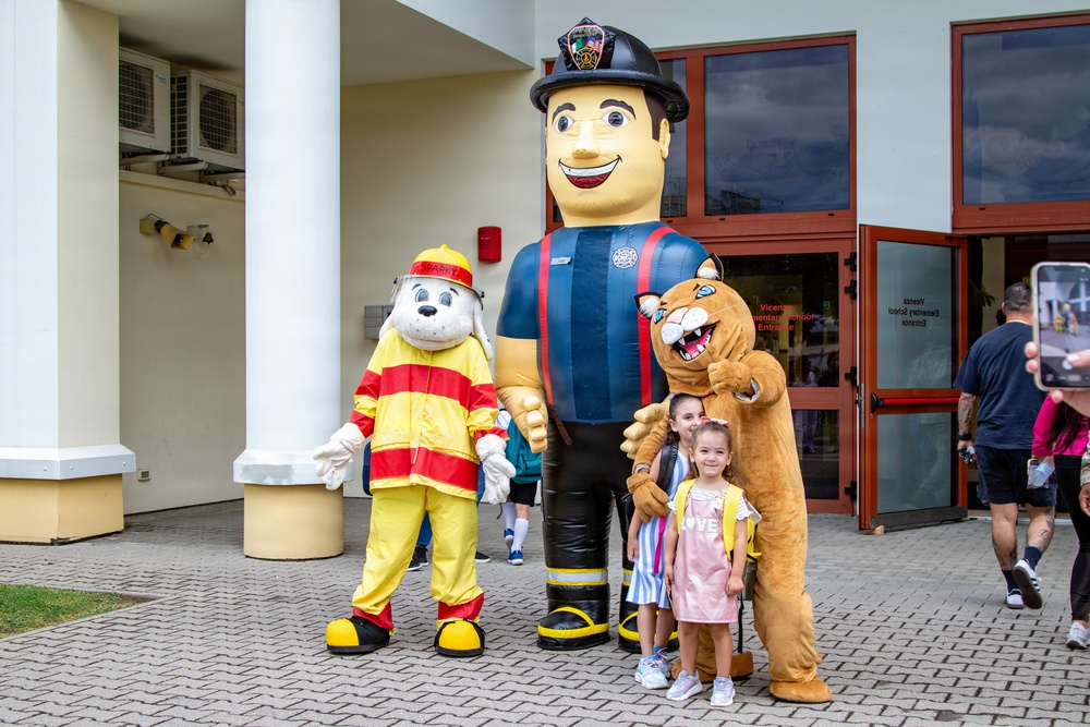 Two children pose for a photo in front of what appears to be a school entrance labeled 'Vicenza Elementary School.' They are flanked by three mascot characters: a dog dressed as a firefighter, a large inflatable firefighter figure, and a lion/cougar mascot. The event appears to be a school welcome or orientation day.