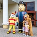 Two children pose for a photo in front of what appears to be a school entrance labeled 'Vicenza Elementary School.' They are flanked by three mascot characters: a dog dressed as a firefighter, a large inflatable firefighter figure, and a lion/cougar mascot. The event appears to be a school welcome or orientation day.