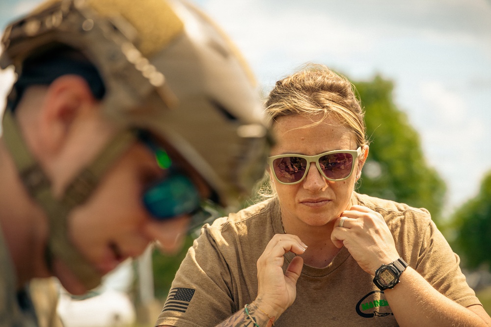 Two individuals are shown in an outdoor setting, one wearing tactical military gear including a tan helmet and green-mirrored sunglasses, and the other a woman in a tan t-shirt with a US flag patch and a digital watch. The woman appears focused and is adjusting something near her collar or chin area. The image has a shallow depth of field with the foreground figure blurred.