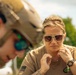 Two individuals are shown in an outdoor setting, one wearing tactical military gear including a tan helmet and green-mirrored sunglasses, and the other a woman in a tan t-shirt with a US flag patch and a digital watch. The woman appears focused and is adjusting something near her collar or chin area. The image has a shallow depth of field with the foreground figure blurred.