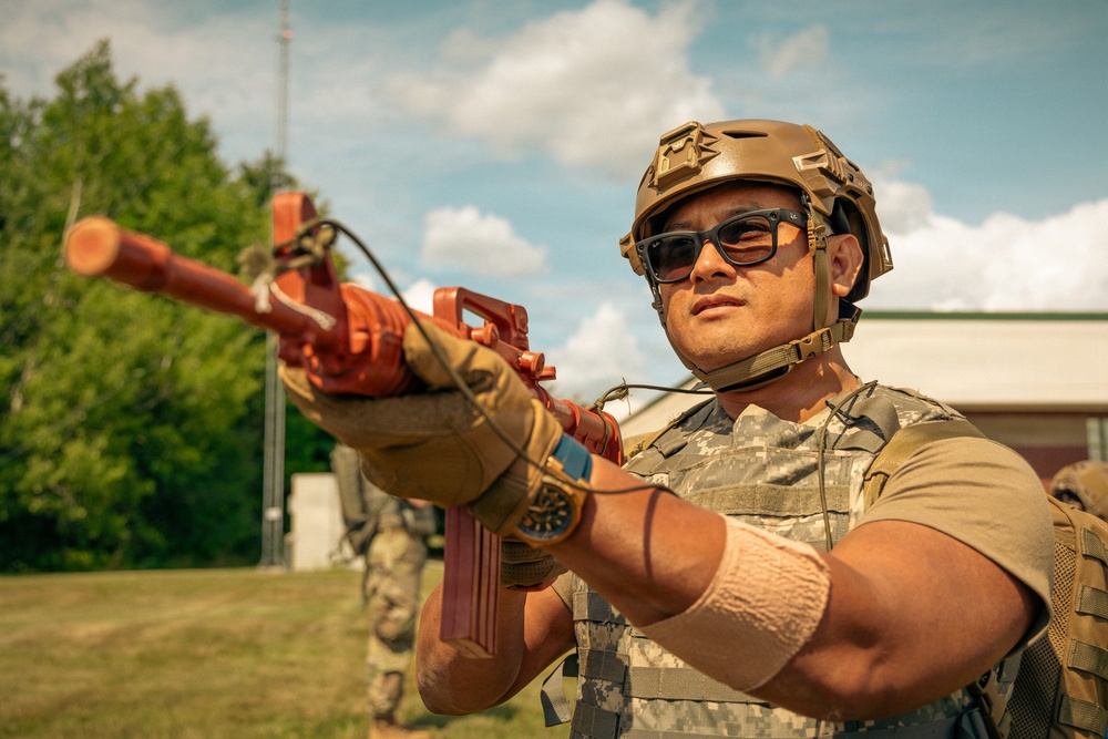 A soldier in full tactical gear is holding a red/orange training weapon (MILES or simunition training device) in an outdoor military training environment. The weapon's distinctive red/orange color indicates it is a training aid rather than a live-fire weapon. Additional soldiers are visible in the background, suggesting a group training exercise.