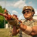 A soldier in full tactical gear is holding a red/orange training weapon (MILES or simunition training device) in an outdoor military training environment. The weapon's distinctive red/orange color indicates it is a training aid rather than a live-fire weapon. Additional soldiers are visible in the background, suggesting a group training exercise.