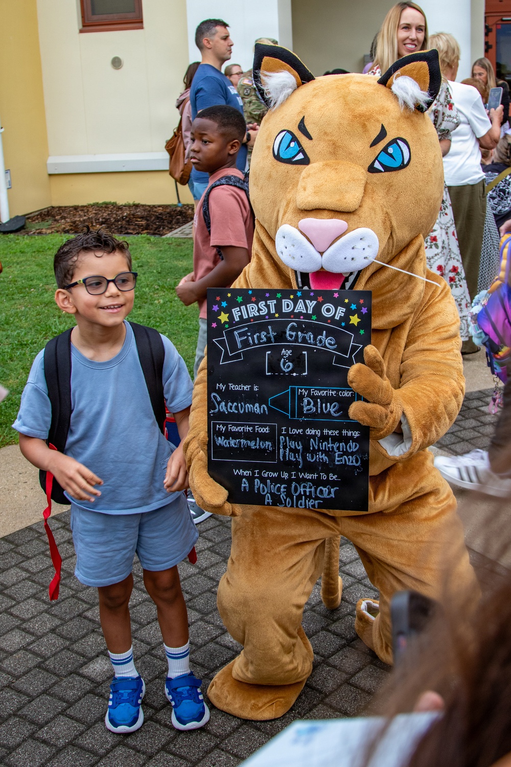 A young boy wearing glasses and a backpack poses next to a school mascot costume (a wildcat/cougar) on his first day of first grade. The mascot is holding a chalkboard sign with the child's personal details including his grade, age, teacher's name, and future aspirations. The scene takes place outside a school building with parents and other students visible in the background.