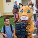 A young boy wearing glasses and a backpack poses next to a school mascot costume (a wildcat/cougar) on his first day of first grade. The mascot is holding a chalkboard sign with the child's personal details including his grade, age, teacher's name, and future aspirations. The scene takes place outside a school building with parents and other students visible in the background.