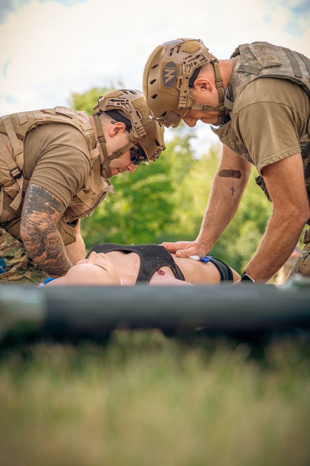 Two military personnel in tactical gear and helmets are leaning over a casualty or training mannequin lying on a stretcher or litter. They appear to be performing tactical combat casualty care (TCCC), assessing or treating a simulated chest wound. The setting is an outdoor grassy area, suggesting a field training exercise.