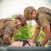 Two military personnel in tactical gear and helmets are leaning over a casualty or training mannequin lying on a stretcher or litter. They appear to be performing tactical combat casualty care (TCCC), assessing or treating a simulated chest wound. The setting is an outdoor grassy area, suggesting a field training exercise.