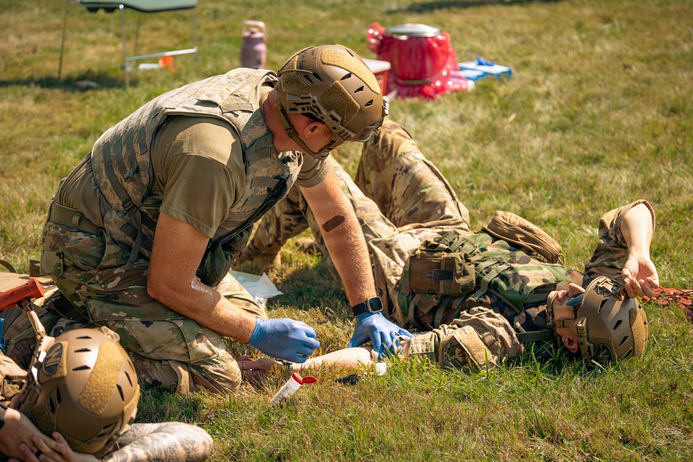 Military personnel are conducting a tactical combat casualty care (TCCC) training exercise in an open grassy field. One soldier wearing blue medical gloves is kneeling and applying what appears to be a tourniquet or bandage to the arm of another soldier lying on the ground. The setting includes training props and equipment visible in the background, indicating a structured field medical training scenario.