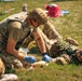 Military personnel are conducting a tactical combat casualty care (TCCC) training exercise in an open grassy field. One soldier wearing blue medical gloves is kneeling and applying what appears to be a tourniquet or bandage to the arm of another soldier lying on the ground. The setting includes training props and equipment visible in the background, indicating a structured field medical training scenario.