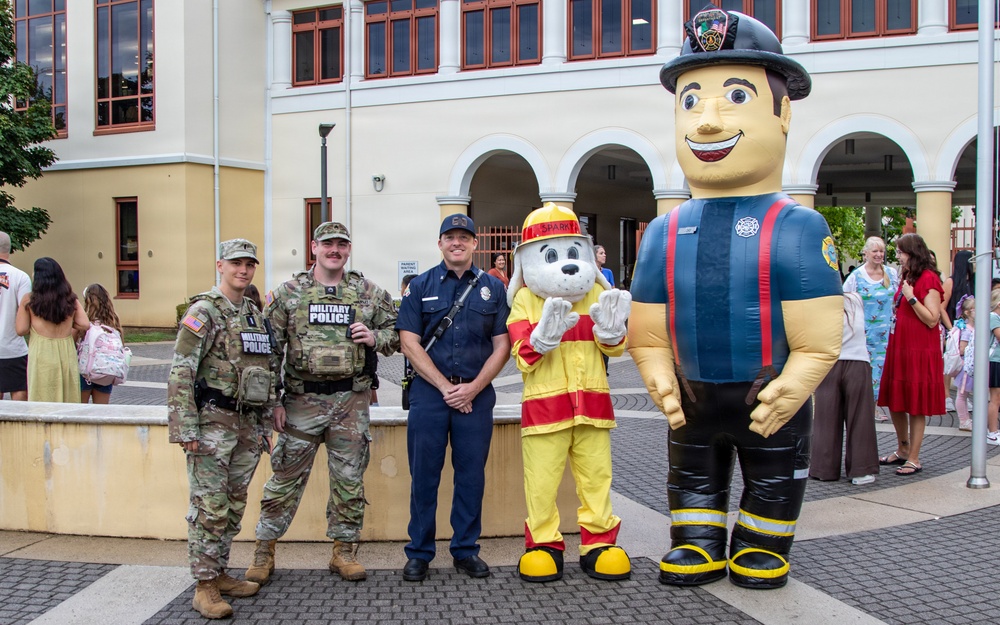 A group photo taken outdoors in front of a large institutional building features two military police soldiers in camouflage uniforms, one civilian law enforcement or fire department officer in blue uniform, a dalmatian fire safety mascot costume, and a large inflatable firefighter character. The setting appears to be a community or school safety event, with families and children visible in the background. The scene suggests a public outreach or back-to-school safety awareness event on what appears to be a military installation.