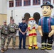 A group photo taken outdoors in front of a large institutional building features two military police soldiers in camouflage uniforms, one civilian law enforcement or fire department officer in blue uniform, a dalmatian fire safety mascot costume, and a large inflatable firefighter character. The setting appears to be a community or school safety event, with families and children visible in the background. The scene suggests a public outreach or back-to-school safety awareness event on what appears to be a military installation.