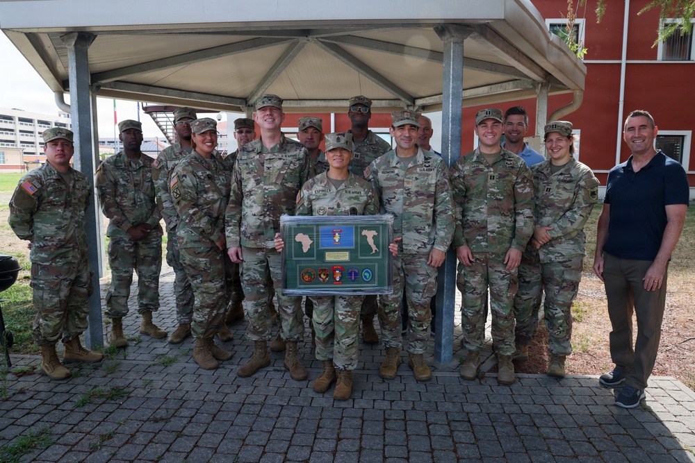 A group of approximately 15 U.S. Army soldiers in OCP (Operational Camouflage Pattern) uniforms pose for a group photo under a covered outdoor area, accompanied by one civilian in a blue shirt. A soldier identified by nametape as 'ESPINOZA' holds a framed shadow box display containing military patches, maps, and what appears to be a commemorative plaque. The setting appears to be a military installation, possibly overseas based on the Italian flag visible in the background.