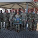 A group of approximately 15 U.S. Army soldiers in OCP (Operational Camouflage Pattern) uniforms pose for a group photo under a covered outdoor area, accompanied by one civilian in a blue shirt. A soldier identified by nametape as 'ESPINOZA' holds a framed shadow box display containing military patches, maps, and what appears to be a commemorative plaque. The setting appears to be a military installation, possibly overseas based on the Italian flag visible in the background.