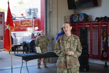 A female U.S. Army officer in OCP (Operational Camouflage Pattern) uniform is speaking at what appears to be an official ceremony inside a fire station at Fort Myer. A red Army general officer flag with a single star is visible to her left, indicating a Brigadier General's presence. Behind her, another soldier in uniform stands near a table with a framed certificate or plaque.