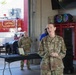A female U.S. Army officer in OCP (Operational Camouflage Pattern) uniform is speaking at what appears to be an official ceremony inside a fire station at Fort Myer. A red Army general officer flag with a single star is visible to her left, indicating a Brigadier General's presence. Behind her, another soldier in uniform stands near a table with a framed certificate or plaque.