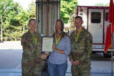 Three individuals pose for a formal photo at what appears to be a military fire station or emergency services facility. Two individuals in U.S. Army camouflage uniforms (OCP pattern) flank a civilian woman who is holding a framed certificate or award. All three are wearing green and gold medallion lanyards, suggesting a recognition or award ceremony.