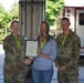 Three individuals pose for a formal photo at what appears to be a military fire station or emergency services facility. Two individuals in U.S. Army camouflage uniforms (OCP pattern) flank a civilian woman who is holding a framed certificate or award. All three are wearing green and gold medallion lanyards, suggesting a recognition or award ceremony.