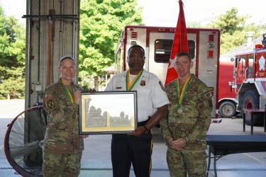 Three individuals are posed together at what appears to be a fire station, with two wearing U.S. Army camouflage uniforms and one wearing a fire department uniform. The center individual, in a fire chief or officer's white uniform shirt, is holding a framed award or plaque. All three are wearing green and gold lanyards or medals, suggesting a formal recognition ceremony.