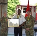 Three individuals are posed together at what appears to be a fire station, with two wearing U.S. Army camouflage uniforms and one wearing a fire department uniform. The center individual, in a fire chief or officer's white uniform shirt, is holding a framed award or plaque. All three are wearing green and gold lanyards or medals, suggesting a formal recognition ceremony.