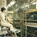 Two workers in white laboratory or industrial coveralls are seated at an industrial machine in what appears to be a manufacturing or processing facility. The setting shows a complex array of pipes, valves, machinery, and a control panel on the left wall. The photograph appears to date from the 1960s or 1970s based on color tone and equipment style.