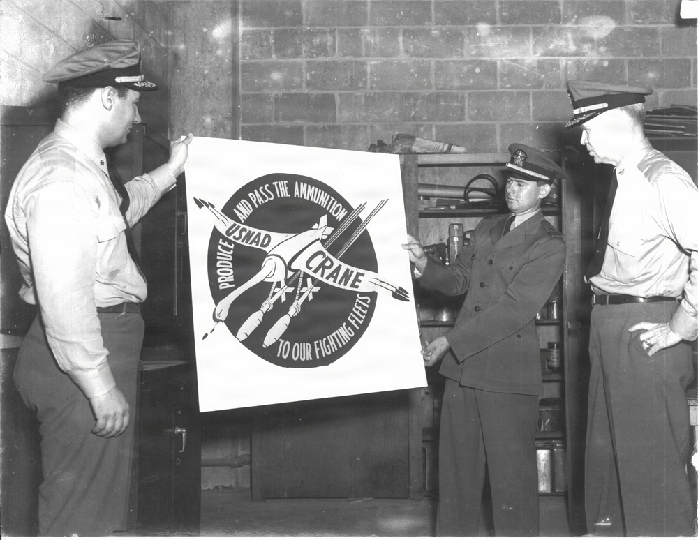 A black-and-white photograph showing three military personnel examining or displaying a promotional poster for USNAD Crane (U.S. Naval Ammunition Depot, Crane). The poster reads 'Produce and Pass the Ammunition to Our Fighting Fleets' with a circular emblem featuring crossed ammunition/weapons. The setting appears to be an indoor facility with a brick wall background, consistent with a WWII-era military installation.
