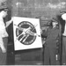 A black-and-white photograph showing three military personnel examining or displaying a promotional poster for USNAD Crane (U.S. Naval Ammunition Depot, Crane). The poster reads 'Produce and Pass the Ammunition to Our Fighting Fleets' with a circular emblem featuring crossed ammunition/weapons. The setting appears to be an indoor facility with a brick wall background, consistent with a WWII-era military installation.