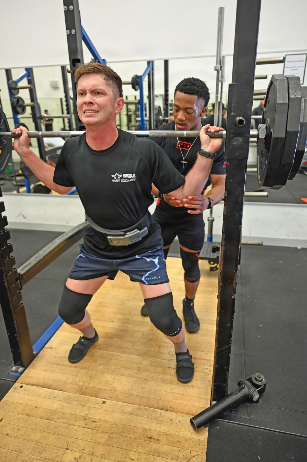 Two men are training in a weightlifting gym, with one man performing a barbell back squat while the other spots him from behind. The lifter is wearing knee sleeves, a lifting belt branded 'SBD', and a black t-shirt reading 'The Beast'. The spotter stands closely behind with hands ready to assist, wearing a black SBD-branded shirt.