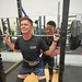 Two men are training in a weightlifting gym, with one man performing a barbell back squat while the other spots him from behind. The lifter is wearing knee sleeves, a lifting belt branded 'SBD', and a black t-shirt reading 'The Beast'. The spotter stands closely behind with hands ready to assist, wearing a black SBD-branded shirt.