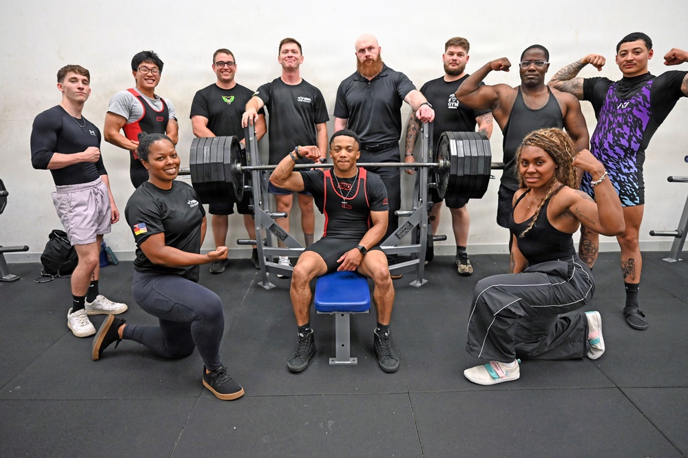 A group of approximately 13 athletes pose together in a gym setting around a loaded barbell on a bench press rack. The group appears to be a powerlifting or strength training team, with several members flexing and posing confidently. The central figure is seated on the bench press bench while teammates flank him in two rows.