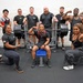 A group of approximately 13 athletes pose together in a gym setting around a loaded barbell on a bench press rack. The group appears to be a powerlifting or strength training team, with several members flexing and posing confidently. The central figure is seated on the bench press bench while teammates flank him in two rows.