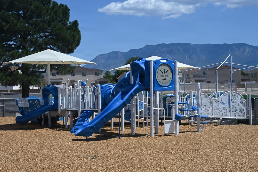 A modern, well-maintained playground featuring blue and white equipment set on a wood chip safety surface. The facility includes multiple slides, climbing structures, and shade canopies, situated in what appears to be a residential neighborhood with a dramatic mountain range visible in the background. The playground appears new or recently installed and is currently unoccupied.