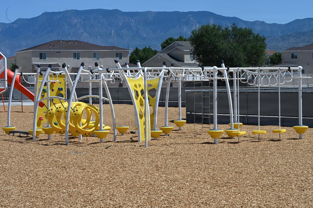 A modern playground featuring yellow and white metal equipment is situated on a wood chip safety surface in a residential neighborhood. The equipment includes climbing structures, monkey bars, and balance elements. Large mountains are visible in the background, suggesting a southwestern U.S. location such as Albuquerque, New Mexico.