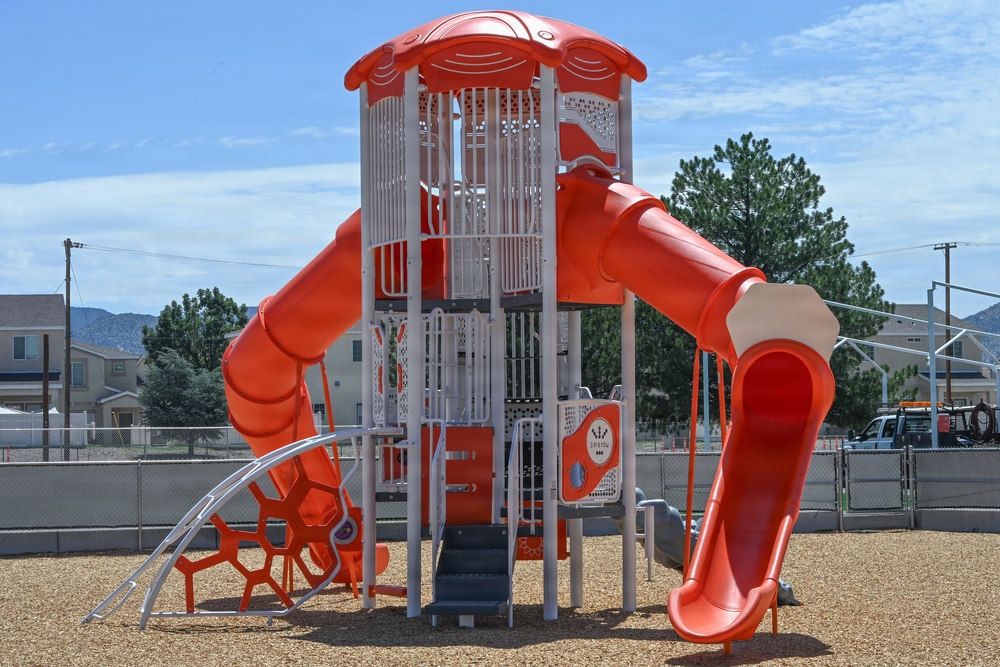 A modern, brightly colored red and white playground structure is situated in a fenced recreational area with wood chip ground cover. The equipment appears new or recently installed, featuring multiple play elements including slides and climbing structures. The setting is in a residential neighborhood with mountains visible in the background under a partly cloudy sky.