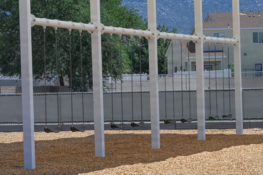 A playground swing set with multiple swings is shown empty during what appears to be a sunny day. The structure features white metal poles and horizontal bars with dark rubber seats suspended by metal chains over a wood chip safety surface. The playground is enclosed by a chain-link fence with residential houses and mountains visible in the background.