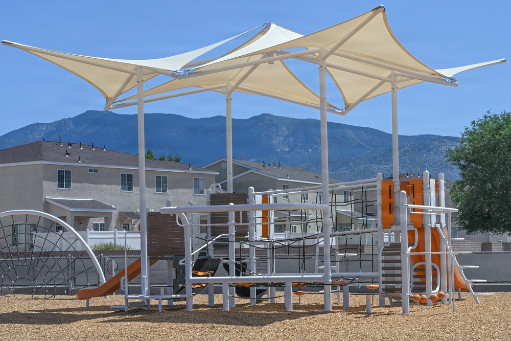A modern residential playground featuring white metal structures with orange accents, covered by large cream-colored tensile shade sails. The equipment is set on a wood chip safety surface within what appears to be a suburban neighborhood community. A mountain range is visible in the background under a clear blue sky.