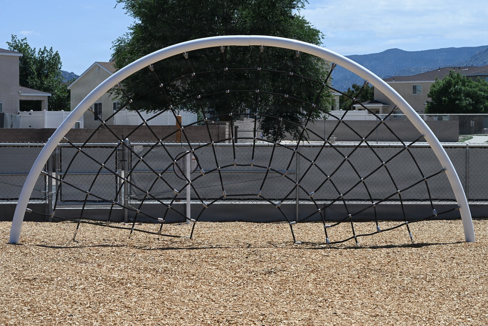 The image shows a dome-shaped rope climbing structure on a playground covered with wood chip mulch safety surfacing. The equipment features a white arched metal frame with a black rope net in a spider-web pattern. The playground is located in what appears to be a residential neighborhood, enclosed by a chain-link fence with mountains visible in the background.