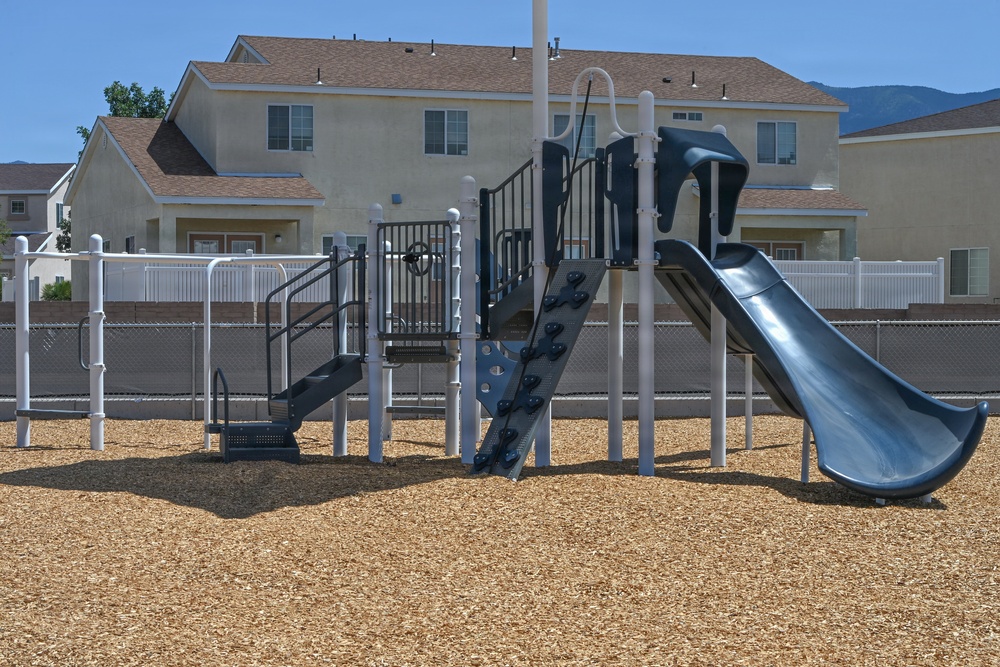 A newly installed playground structure is situated in a residential neighborhood, surrounded by wood chip ground cover. The equipment features a slide, climbing wall, stairs, and railings in a dark gray and white color scheme. The area appears clean and unused, suggesting recent installation.
